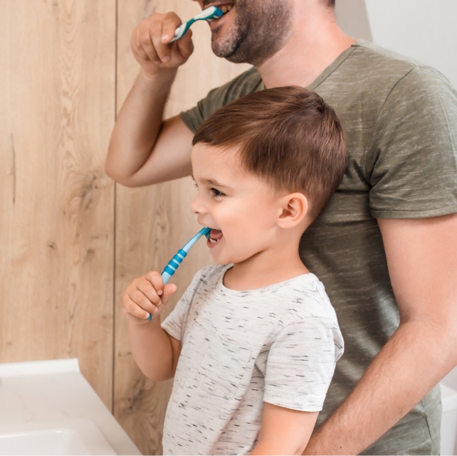 Small child brushing their teeth with their father, which is important after a visit to the dentist